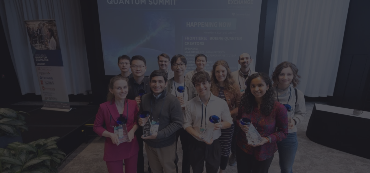 A group of 12 award recipients poses at the Chicago Quantum Summit, holding clear trophies with blue accents. The background features a large screen with the event title and a banner of member organizations.