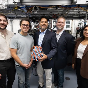Five people in a laboratory setting with electronic equipment, one holding a geometric object.