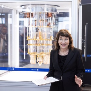 A woman stands in front of a glass case containing a complex gold-colored apparatus, likely a quantum computer, with &quot;UNITED&quot; barriers around them.