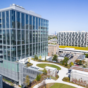 A modern multi-story building with a glass facade, surrounded by landscaped areas, city buildings, and a distant skyline under a blue sky.