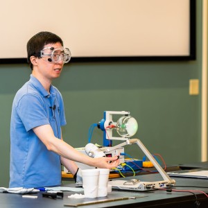 A man in googles stands at a desk behind equipment as he does a demonstration at the orientiation.