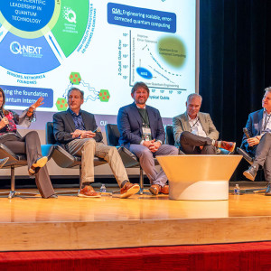 Five panelists seated on stage in front of a screen displaying quantum technology graphics.