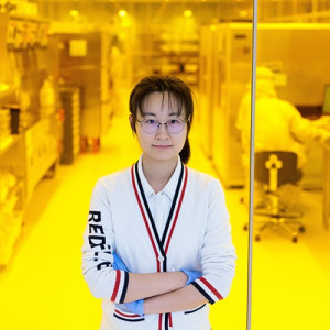 PhD student Zixi Li stands in front of a glass walling showing a yellow-tinged lab, with shelves and equipment in the background.