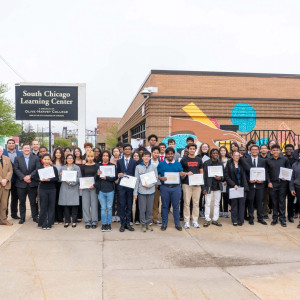 the 2026 cohort of high school students graduated from Fermilab's Saturday Morning Quantum program stand outside a brick building holding certificates.