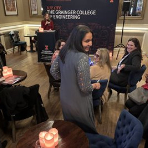 Illinois Physics Professor Smitha Vishveshwara beams while American Institute of Physics Chief of Staff Liza Dart Caron (standing behind a podium) speaks about Vishveshwara's public engagement efforts at Chicago's Den Theater.