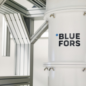 A white cylindrical dilution refrigerator with the Bluefors logo