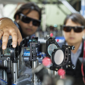 Two researchers wearing laser safety glasses adjust optical components on a photonics lab bench. One reaches toward a beam-splitting optic mounted on a precision post; mirrors, lenses, and fiber optic cables are arranged across the optical breadboard in the foreground.