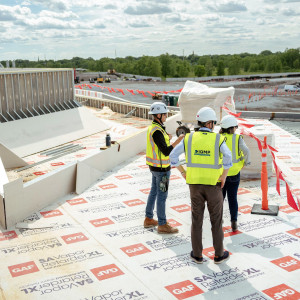 Three people in hard hats and safety vests stand on a building rooftop overlooking the PsiQuantum site.