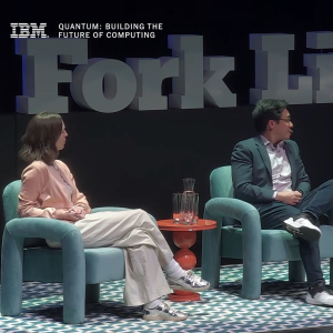 Two people seated in conversation on a stage with teal chairs and orange table.