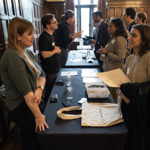 A networking event with attendees engaged in conversation around a table covered with a black cloth. Visible items include water bottles, papers, and a tote bag labeled "SANDBOX."