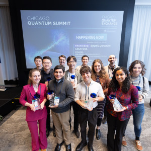 A group of 12 award recipients poses at the Chicago Quantum Summit, holding clear trophies with blue accents. The background features a large screen with the event title and a banner of member organizations.