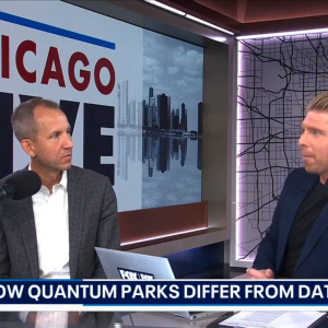 Two men discuss topics on a talk show set with a backdrop displaying "CHICAGO LIVE" and a skyline reflection. One man is in a gray blazer, while the other wears a dark suit.