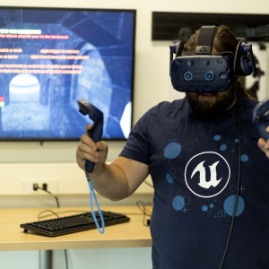 Person wearing a VIVE virtual-reality headset and holding two motion controllers in front of a monitor displaying a virtual stone corridor.